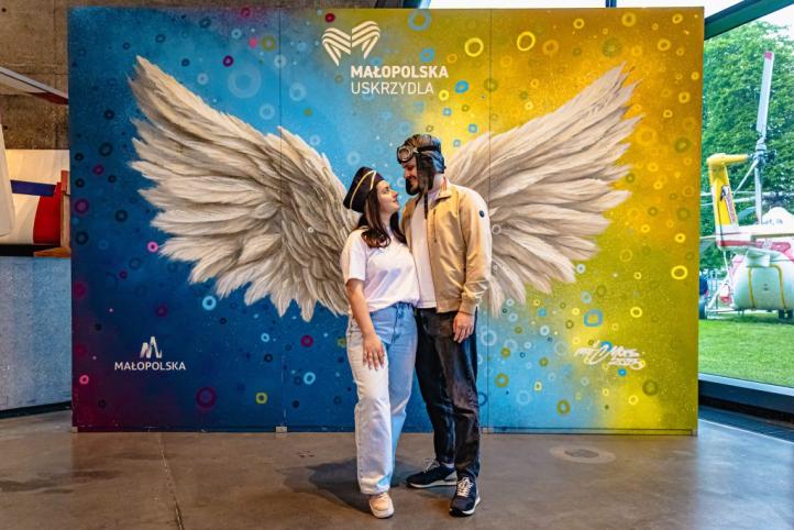 Kobieta i mężczyzna w lotniczych nakryciach głowy na tle baneru ze skrzydłami i logotypem Małopolski (fot. Muzeum Lotnictwa Polskiego) Kobieta i mężczyzna w lotniczych nakryciach głowy na tle baneru ze skrzydłami i logotypem Małopolski (fot. Muzeum Lotnictwa Polskiego)